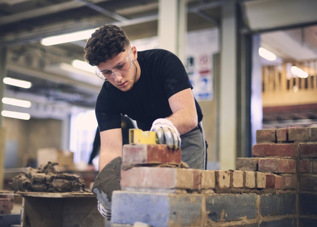 This image shows a student in a bricklaying workshop at Chichester College. They are wearing gloves and safety goggles, and they are laying cement onto a small brick wall that they are constructing. You can see other parts of the workshop in the background too.
