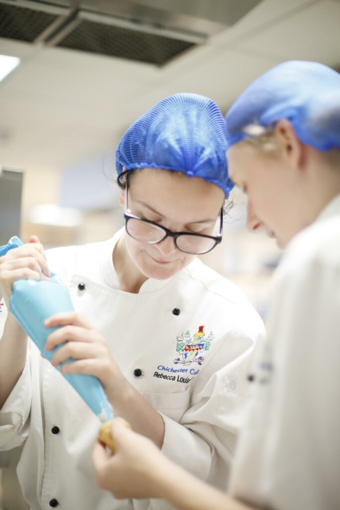 This images shows 2 students in the Catering & hospitality sector using a piping bag. They are in their chef's outfits with hair nets too.