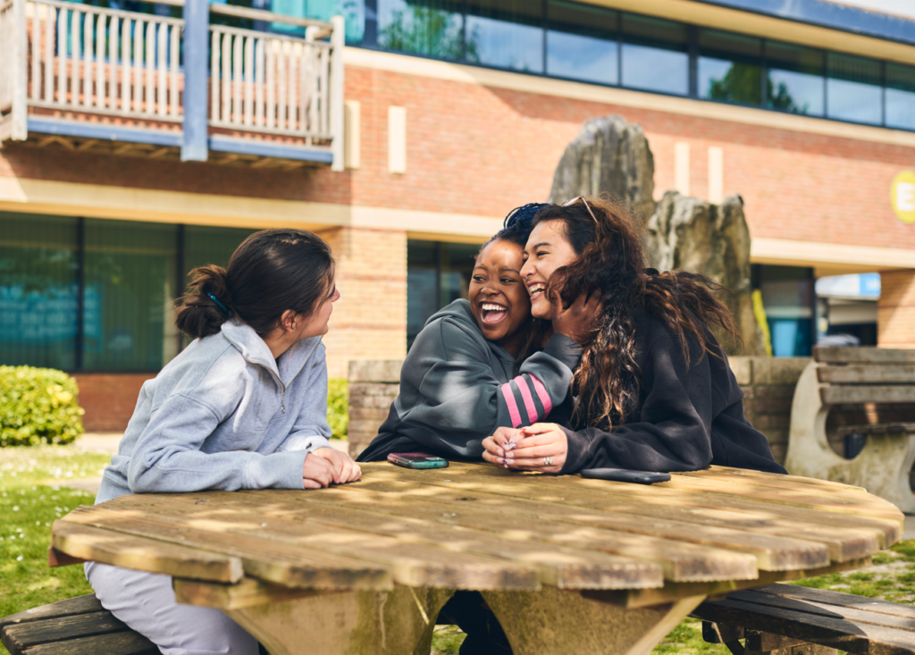 Students smiling and socialising at a bench outside the college